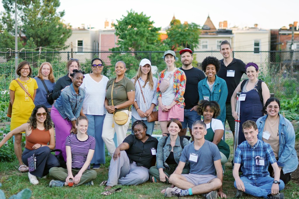 Group of 20 people posing together in front of a garden in an urban setting. 